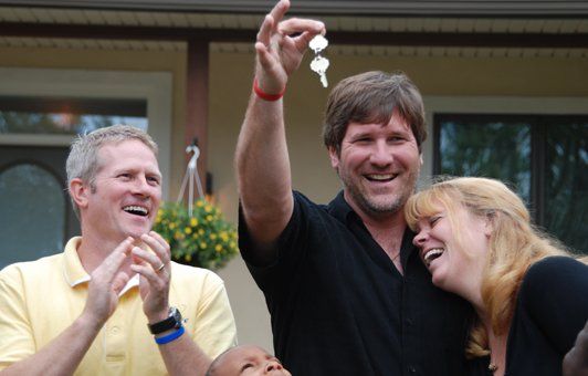 Man holding keys, smiling, with arm raised, celebrates purchase of house with happy couple and man in yellow shirt.