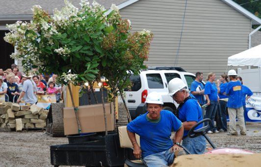 Workers in blue shirts and white hard hats moving plants on a utility vehicle at an outdoor event.