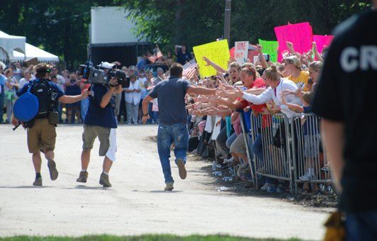Man runs toward a crowd of people behind a barrier, pursued by cameramen. People hold signs.