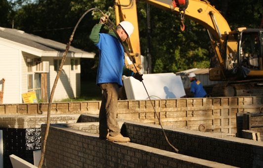 Construction worker using a tool at a building site with an excavator and building materials.
