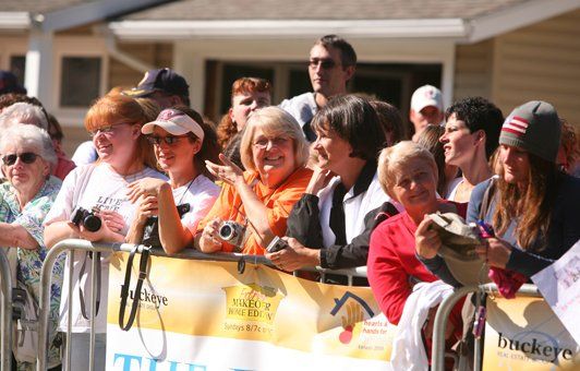 Spectators watch an event behind a barrier, some holding cameras, smiling, and pointing.