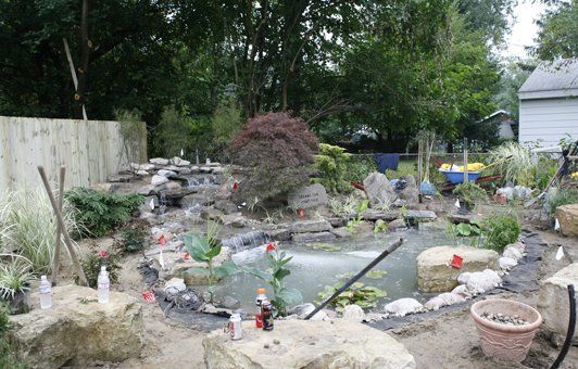 Backyard pond with waterfall, plants, rocks, and a light-colored fence in the background.