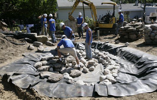 Landscapers building a pond, using rocks and an excavator. They wear blue shirts and white helmets, working outdoors.