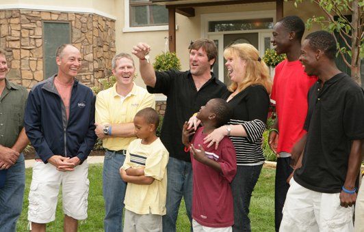 Group of people outside a house, some smiling. A man points upward while looking at a small boy.