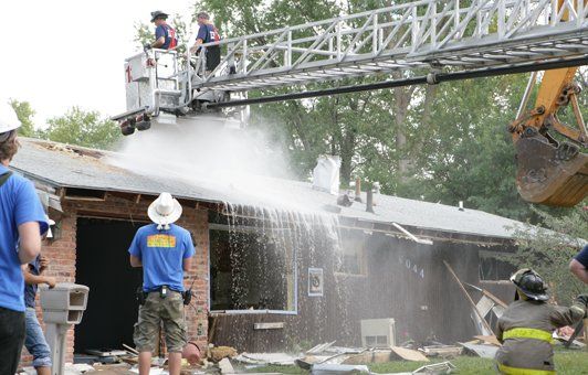 Firefighters on ladder spraying water onto burning house.