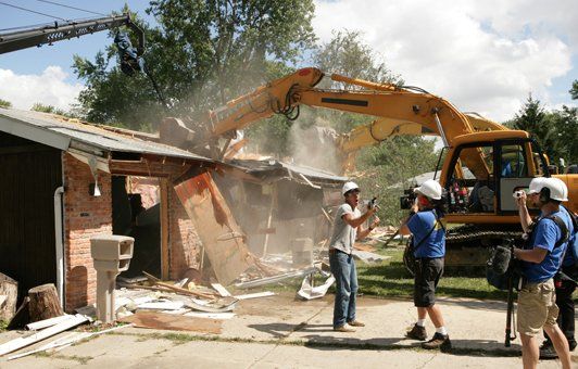Construction crew demolishing a brick building with a large excavator, filmed by a TV crew outdoors.