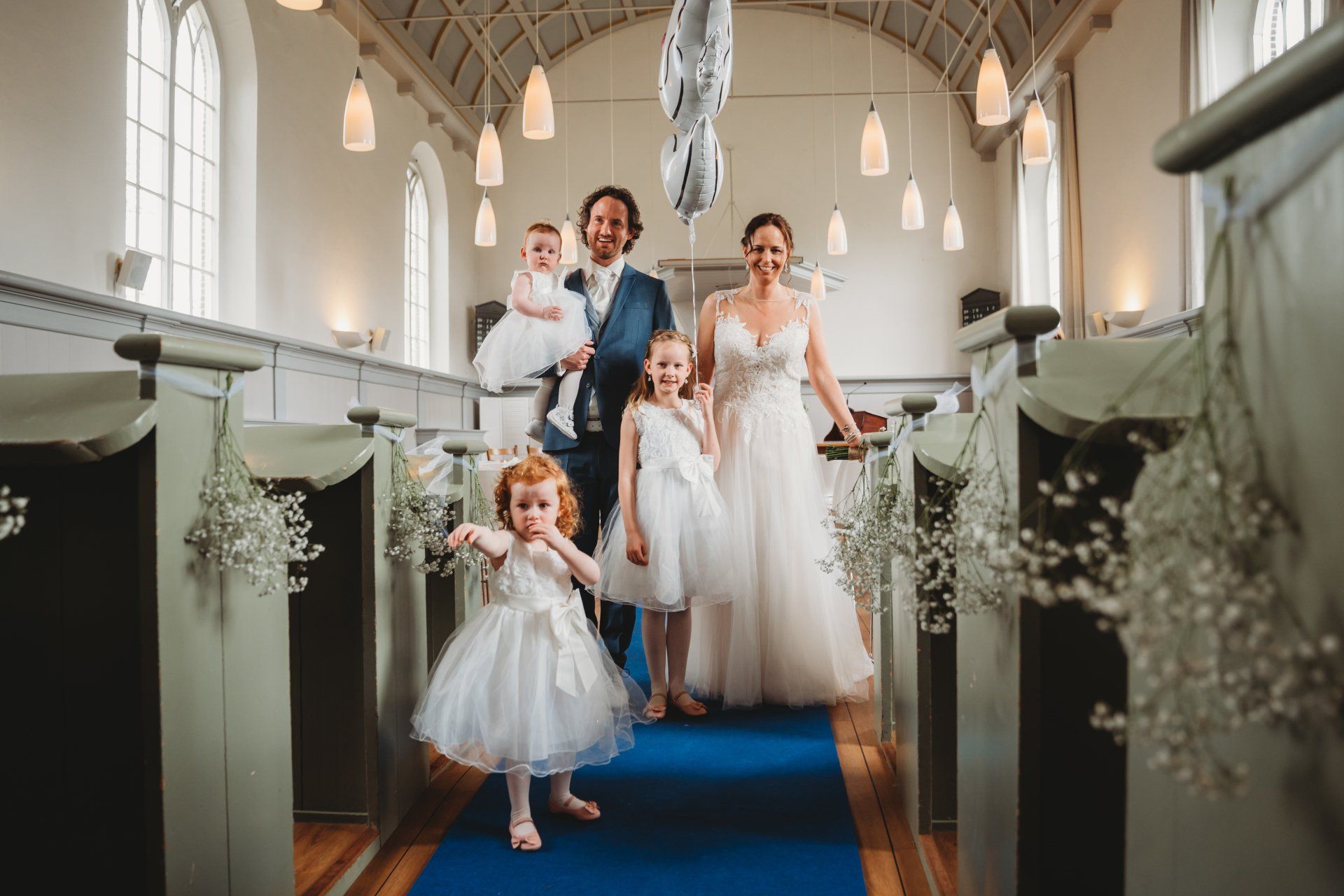 Een bruid en bruidegom lopen met hun bloemenmeisjes door het gangpad van een kerk.