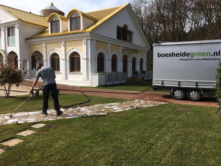 Man wast een stenen pad met een hogedrukreiniger naast een groot huis, vrachtwagen boesheidegreen.nl.