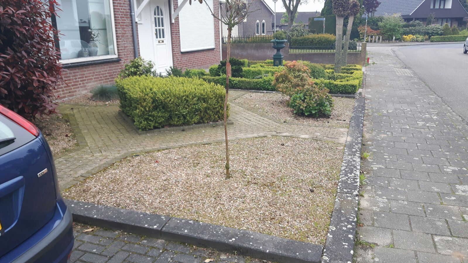 Een voortuin met struiken, stenen en een stenen pad naast een straat en een huis.