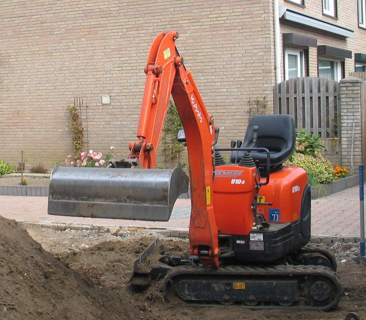 Een oranje minigraafmachine graaft een sleuf op een verhard terrein vlakbij een bakstenen gebouw.