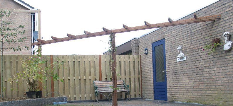 Houten pergola bevestigd aan een bakstenen muur met een blauwe deur. Houten hek en bank zichtbaar.