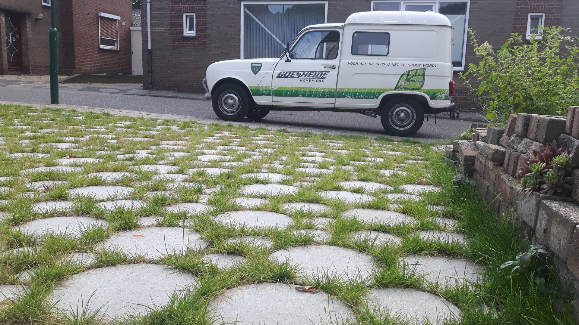 Witte bestelwagen met groen grasdesign geparkeerd op met gras bedekte bestrating. Bakstenen muur en huis zichtbaar.