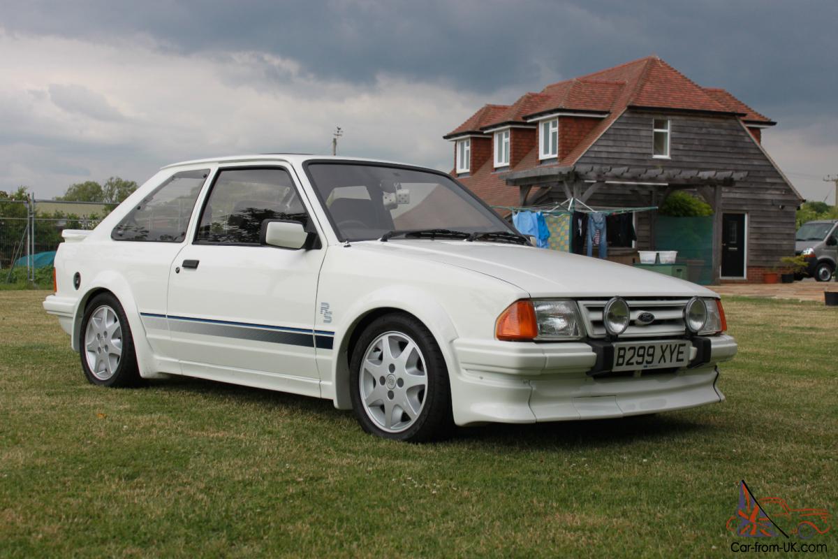 A white car is parked in a grassy field in front of a house.