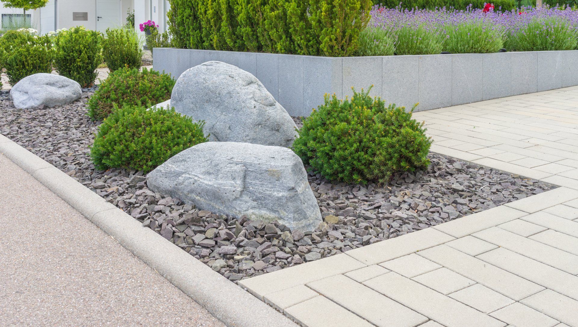 Landscaped gravel bed with large gray boulders and green shrubs beside a paved walkway