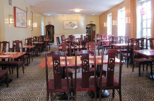Interior of a Chinese restaurant; empty tables with red chairs, carpeted floor, and framed art.