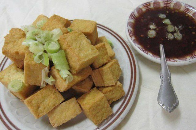 Fried tofu cubes with sliced scallions on a plate, with a dipping sauce and a spoon.