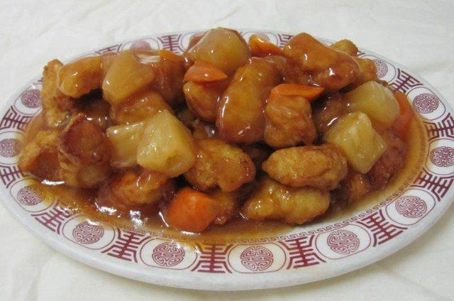 Sweet and sour chicken dish on a white plate with red patterned rim. Chunks of chicken, pineapple, and carrot in sauce.