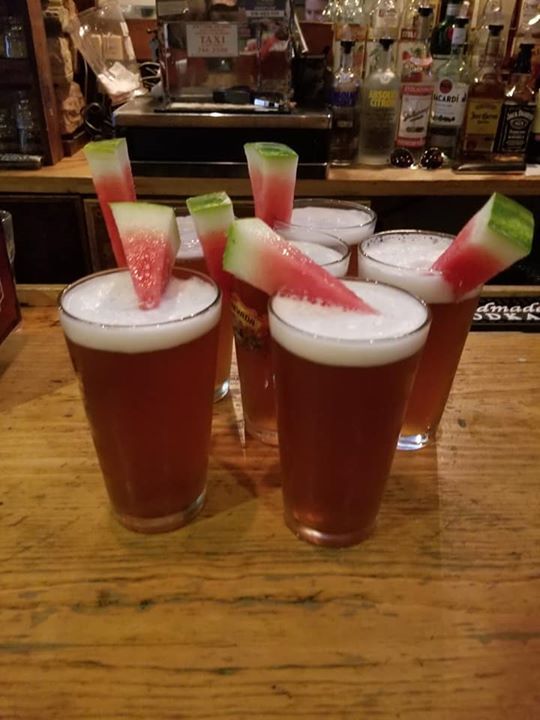 Four glasses of beer with watermelon slices in them on a wooden table.