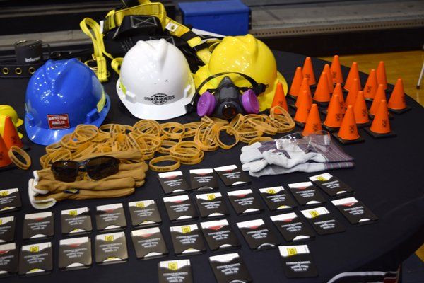 Safety equipment display: blue, white, yellow hard hats, orange cones, sunglasses, gloves, and cards on a black table.