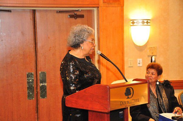 Woman speaking at a podium with a DoubleTree logo. Another woman sits to her right, both indoors.