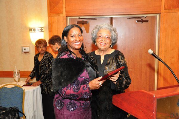 Woman presents award to another woman at a podium; formal setting, smiling.