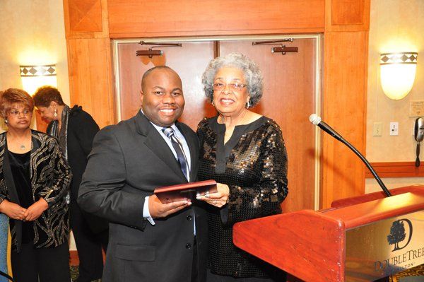 Man in suit receives award from woman in black sequined top. Wooden podium in background.