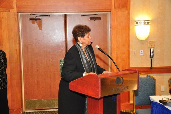 Woman speaking at a podium with a microphone in a room with wood doors and a wall sconce.