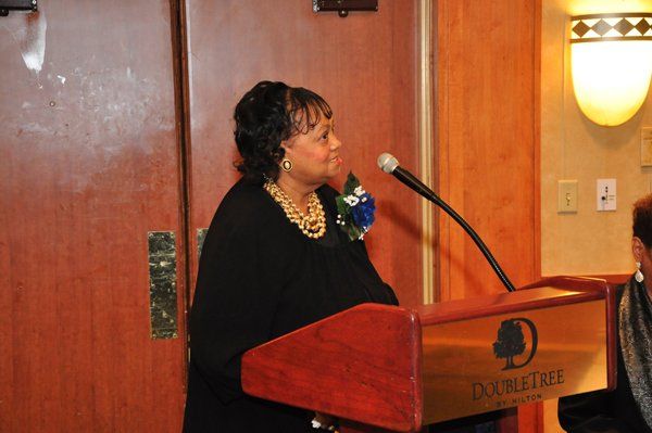 Woman speaking at a podium with a DoubleTree logo, wearing a black top and pearls.