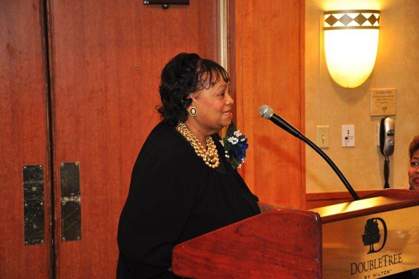 Woman speaking at a podium in a DoubleTree hotel. She wears a black top and necklace.