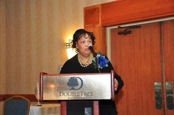 Woman speaking at a DoubleTree podium, wearing a black outfit and a necklace, in a banquet hall.