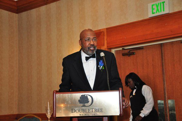 Man speaking at a DoubleTree podium, wearing a tuxedo. A woman stands nearby.