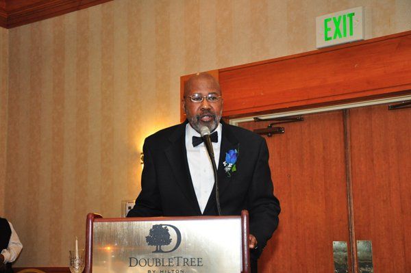 Man in tuxedo speaking at podium, DoubleTree hotel backdrop.
