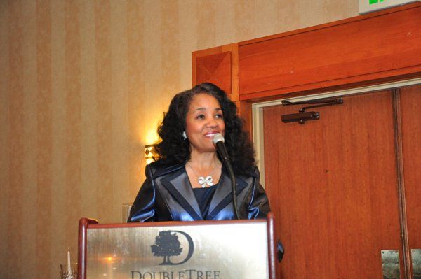 Woman speaking at a DoubleTree hotel podium. She wears a black jacket and smiles.