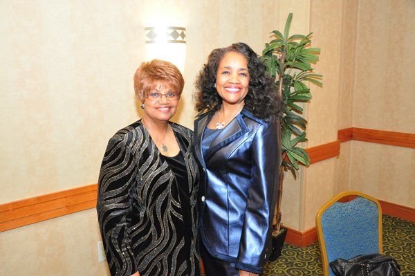 Two women pose in formal attire, smiling against a neutral wall with decorative accents and a plant.