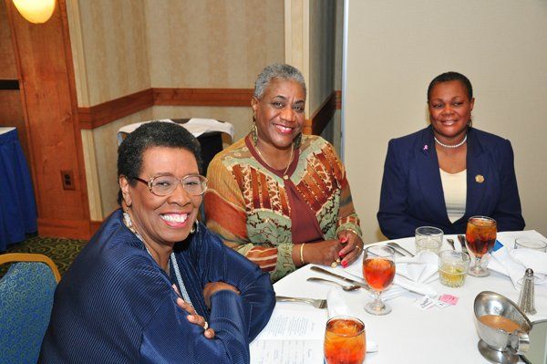 Three women smiling at a table set for a meal. They are indoors with white table linens and drinks.