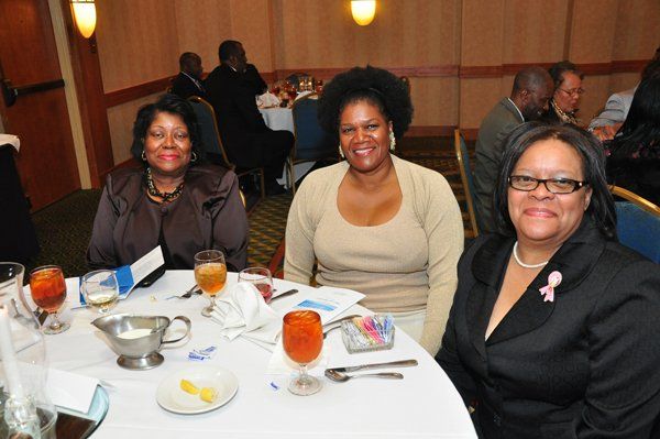 Three women seated at a table in a restaurant; one wears a pink ribbon, cocktails present.