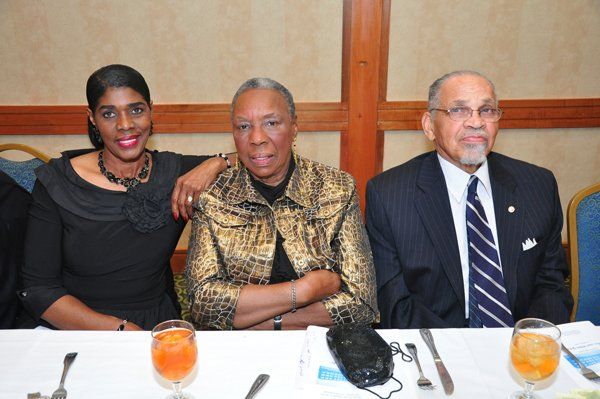 Three people at a table; woman in black arm on woman in gold jacket, man in suit on the right.