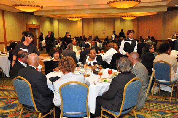 Large banquet hall with round tables and people dining. Waitstaff serves guests.