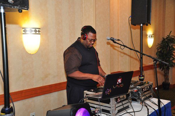 DJ at a party, working on equipment. Dark clothing, headphones, and laptop. Two speakers and microphones nearby.