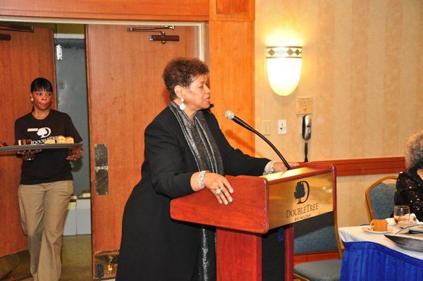 Woman speaking at a podium in front of a “DoubleTree” sign. Server with tray walks by.