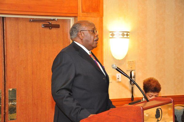 Man in suit speaking at a podium, in a room with a light fixture and a woman partially visible.