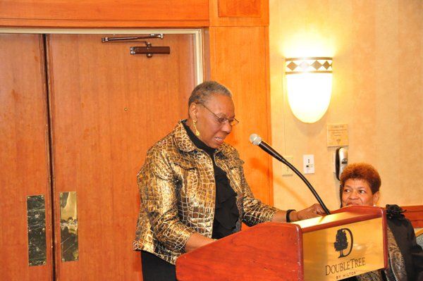 Woman speaking at a DoubleTree hotel podium, wearing a patterned jacket, another woman beside her.