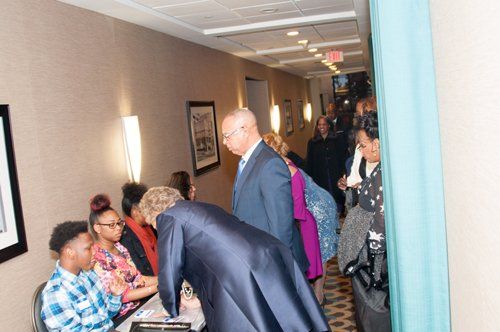 People gathered in a hallway; some appear to be checking in at a desk. Dimly lit; walls are light beige.