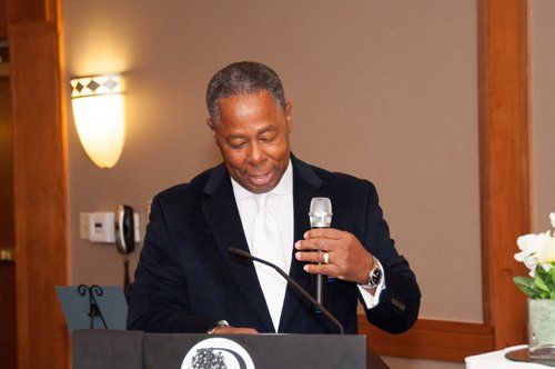 Man in suit speaking at a podium, holding a microphone indoors.