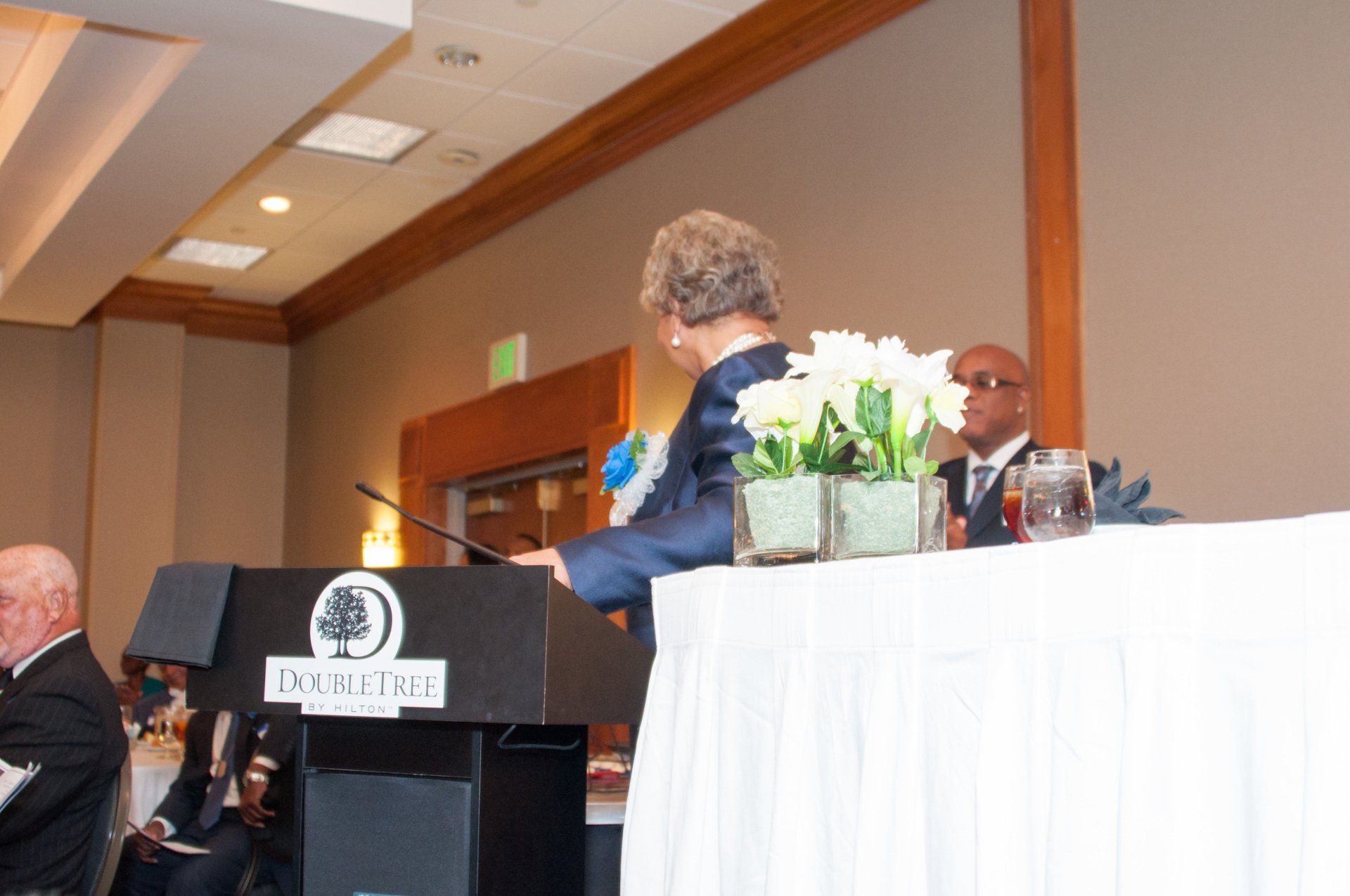 Person at a podium, speaking at a DoubleTree event. Flowers on the table. Other people seated.