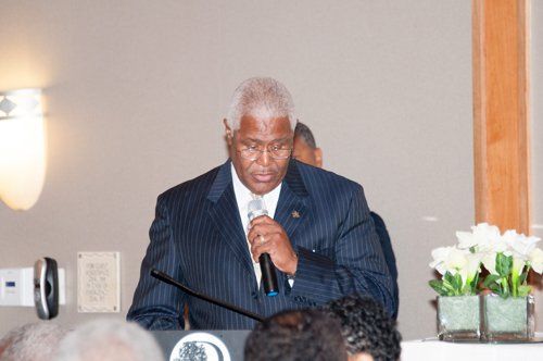 Man in suit speaking at a podium, holding a microphone. Flowers are visible.