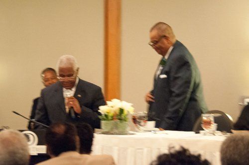 Men in suits at a table, one speaking into a microphone, others nearby.