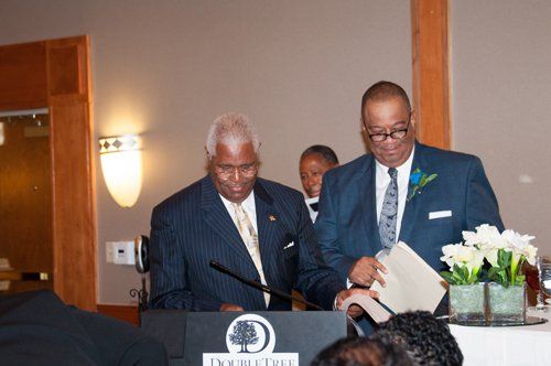 Two men at a podium; one speaks into a microphone, the other holds documents; interior with floral arrangement.