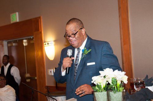 Man in a blue suit speaks into a microphone at an event, near floral arrangement.