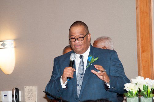 Man in blue suit speaks into microphone, gesturing. Other people visible in the background.